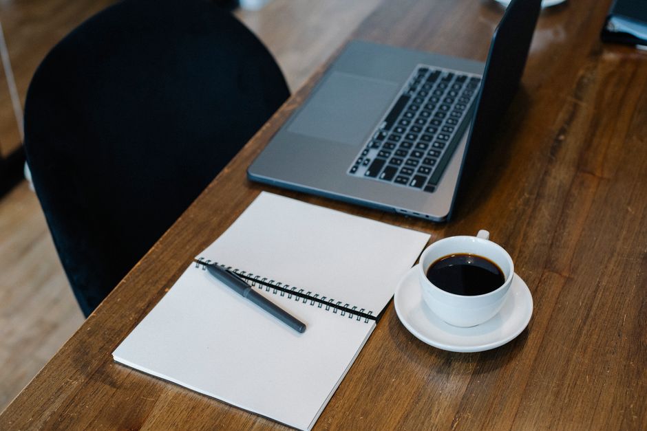an open notebook with a pen, a cup of coffee, and a laptop on a wooden table