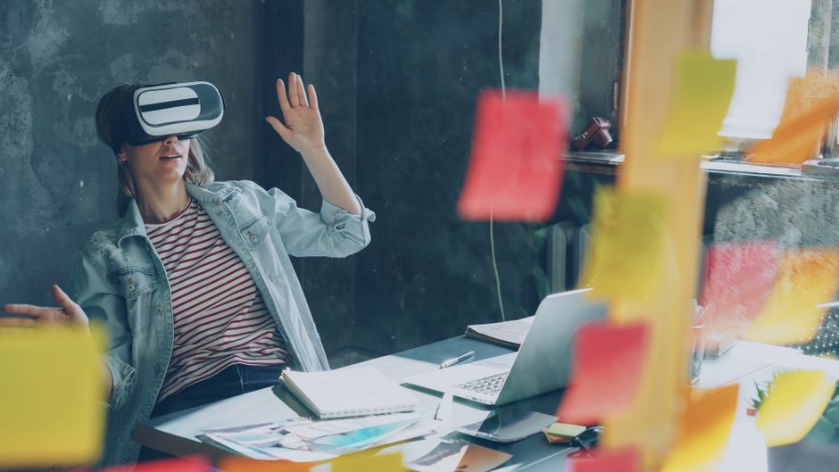 Woman using VR headset at desk, brainstorming with sticky notes and laptop.