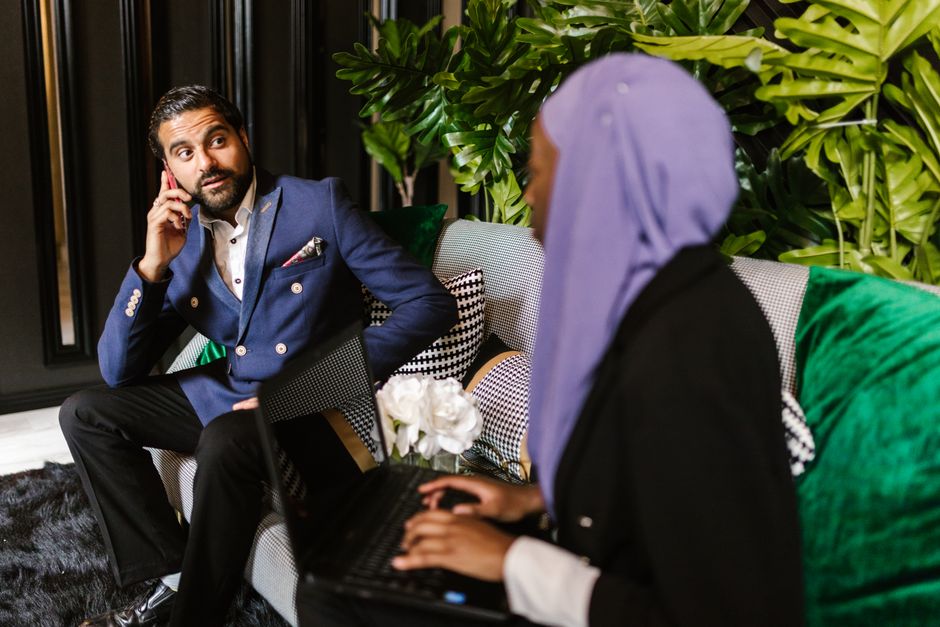 Two colleagues engaged in a business meeting indoors, discussing over a laptop and phone.