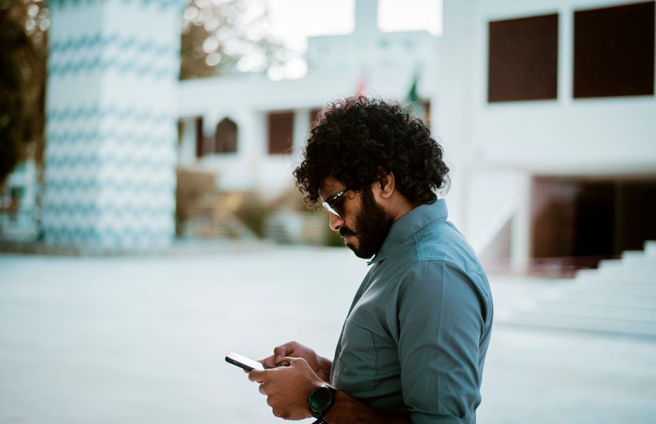 Side view of a curly-haired man with sunglasses using a smartphone in a sunny outdoor setting.