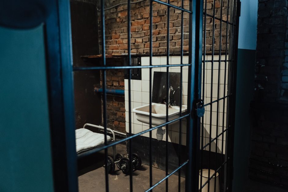 A dimly lit prison cell interior featuring a bed, sink, and exposed brick walls, evoking feelings of isolation.