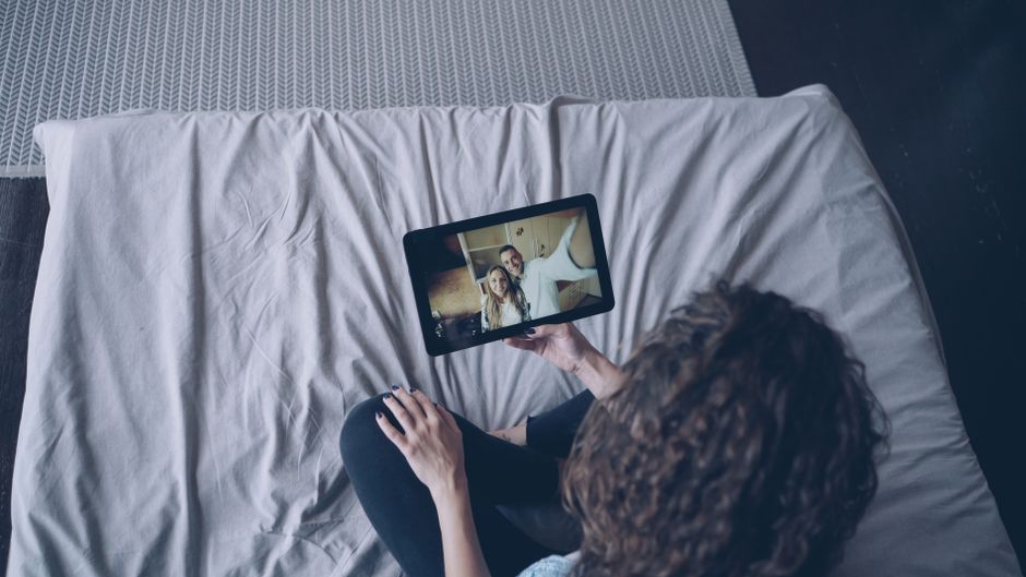 Overhead view of a woman using a tablet for a video call while sitting on a bed.