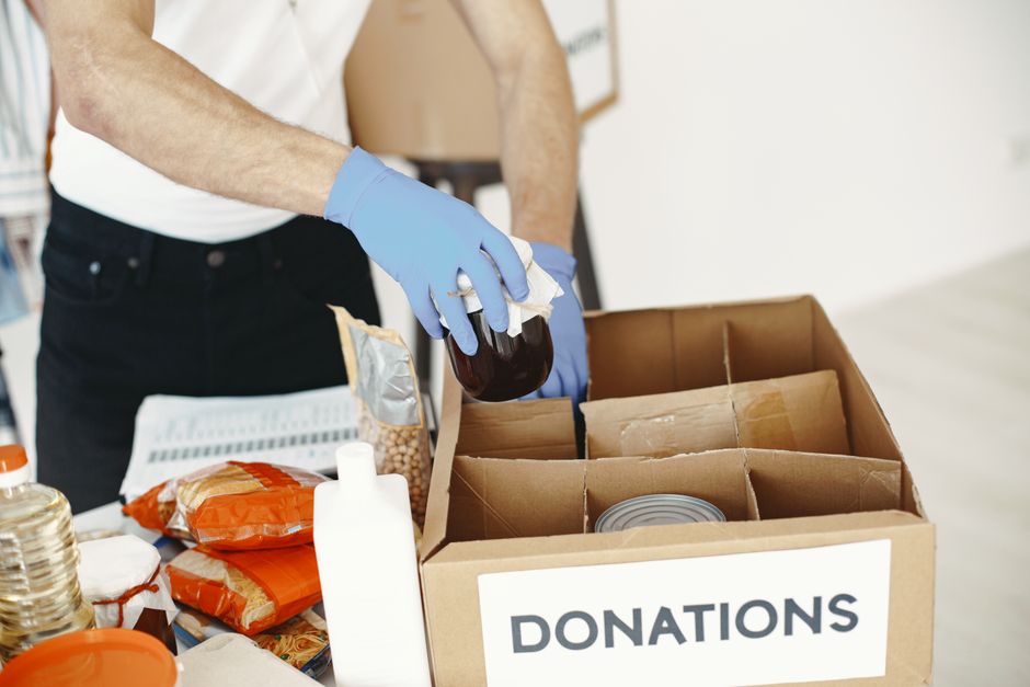 A volunteer with gloves packs canned goods and supplies in a donation box.