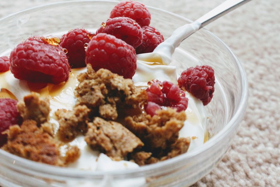 From above closeup of breakfast with tasty natural yogurt decorated with crumbled biscuit and whole ripe raspberries with caramel syrup in glass bowl with spoon on soft plaid in morning