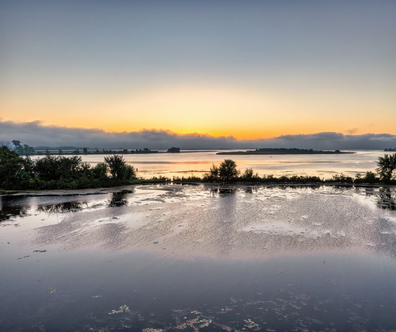 Peaceful sunrise over a calm lake with reflective waters and lush trees, offering a serene landscape view.
