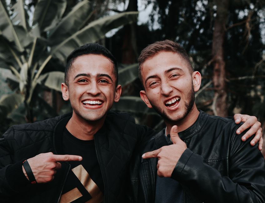 Two young men smiling and pointing at each other in an outdoor setting with lush greenery.