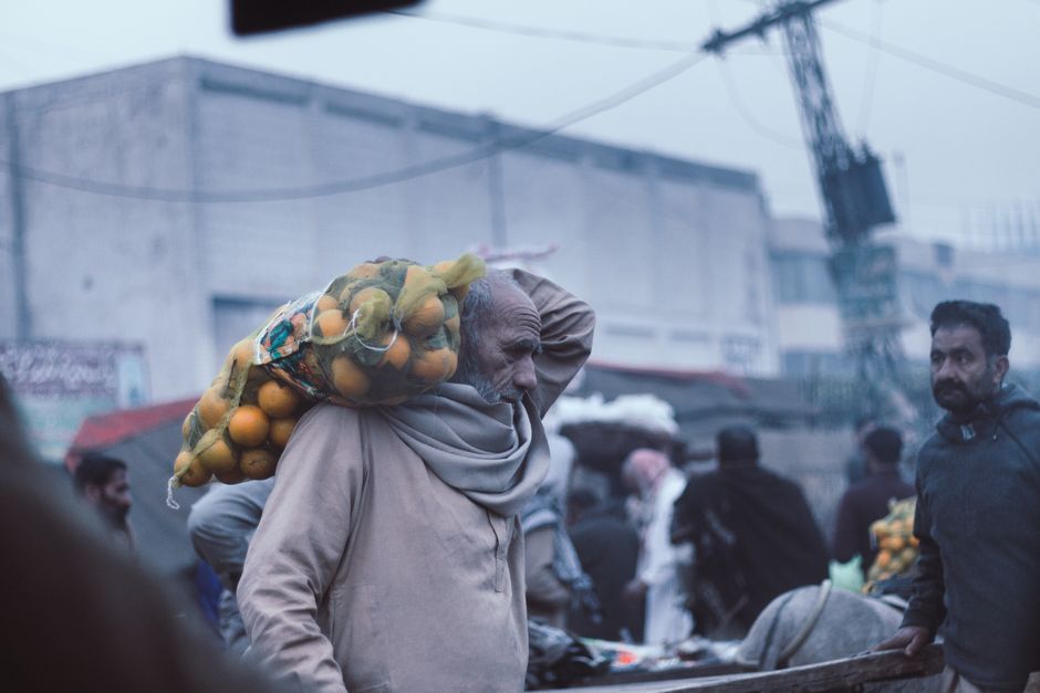 An elderly man carrying oranges through a bustling Lahore market street, showcasing daily life.