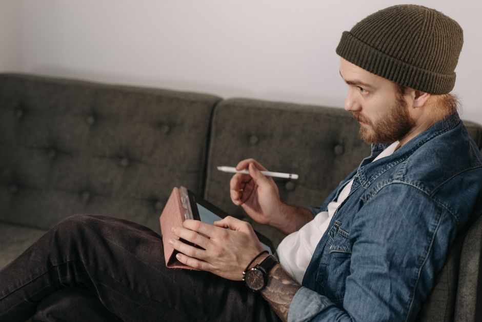 Bearded man in denim jacket sketching on a tablet using a stylus, seated indoors.