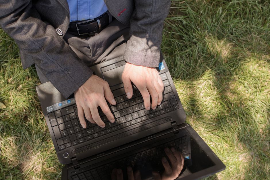 Man working on laptop in a suit sitting on grass, outdoors in daytime.