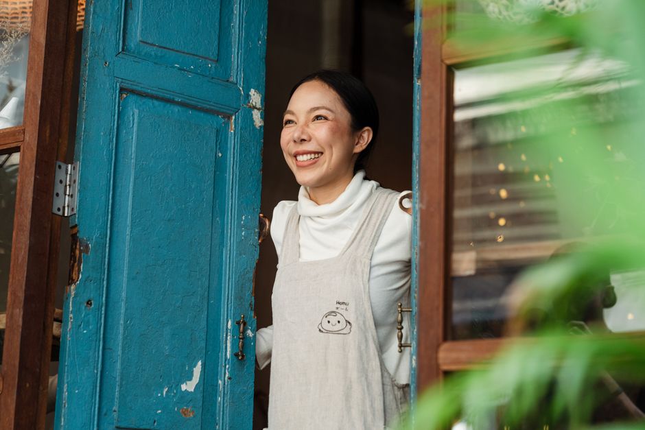 Cheerful ethnic female standing near old blue door with blots on surface and window near plants in daylight and looking away