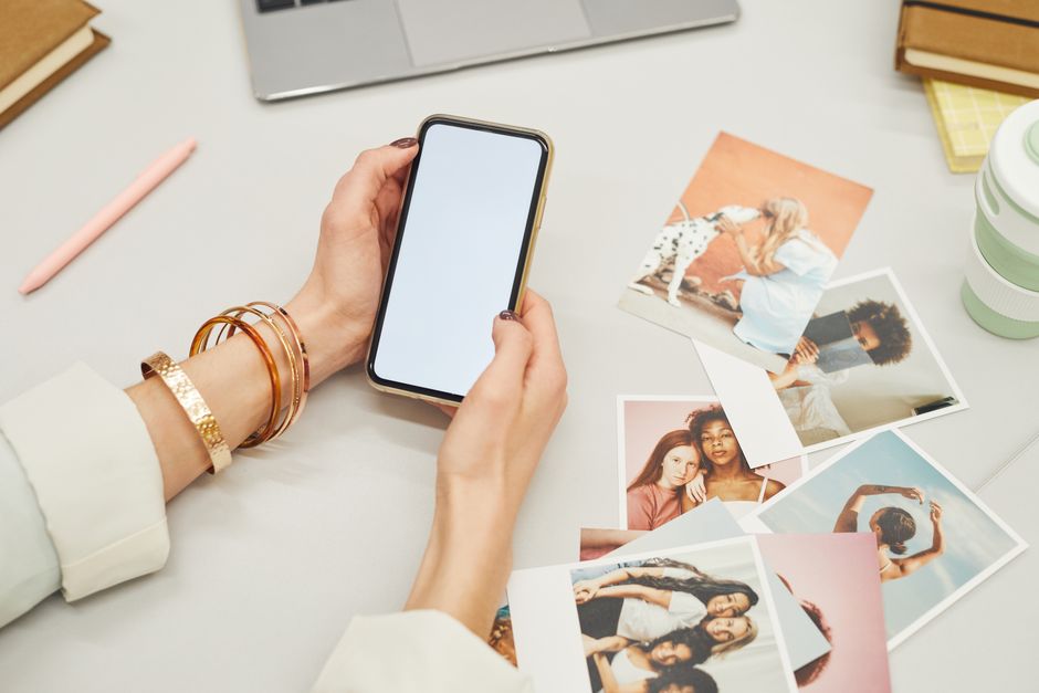 A person holding a smartphone surrounded by printed photographs and a laptop on a desk.