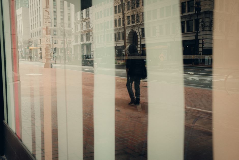 Reflection of a person walking on city streets, seen through a window creating a layered urban view.