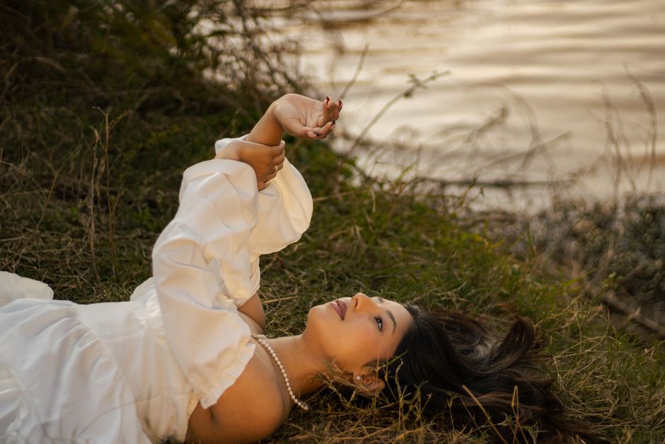 A young woman in a white dress relaxes on grass by the water at sunset, exuding peace.