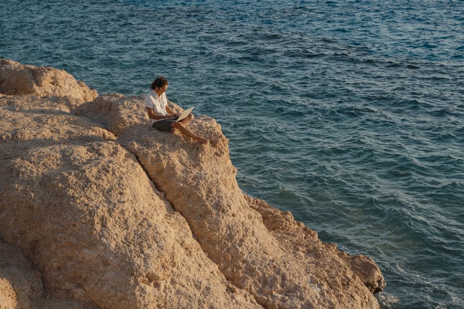 A man sits on a rocky cliff by the sea, working remotely on a laptop.