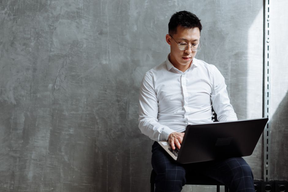 Asian man wearing glasses focused on laptop work. Indoor office setting.