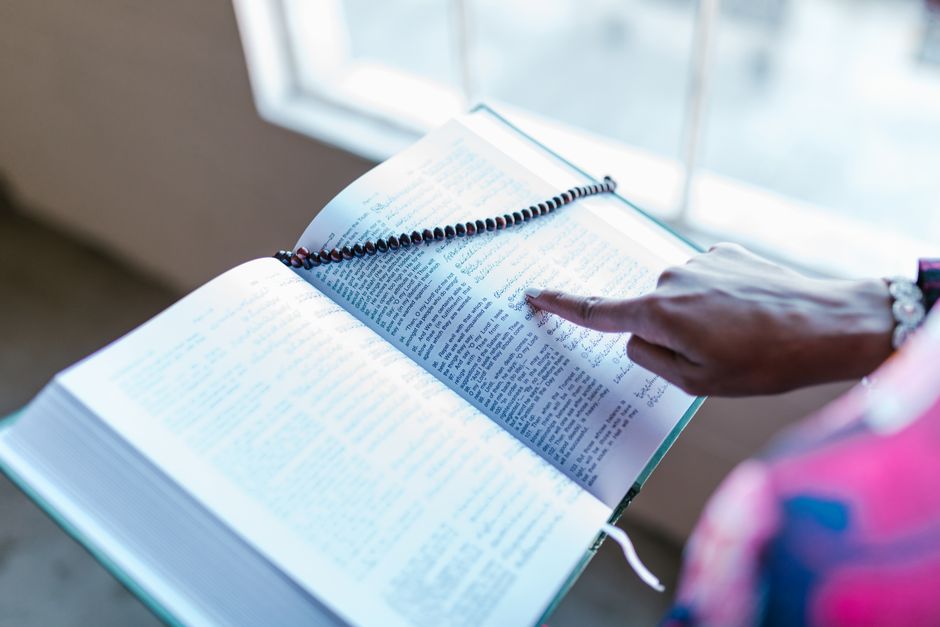A Muslim woman pointing to a passage in the Quran with prayer beads draped over it indoors.
