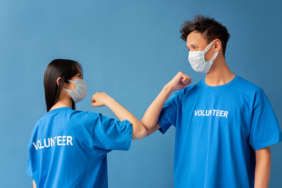 Two volunteers wearing blue shirts and face masks elbow bump for safety.