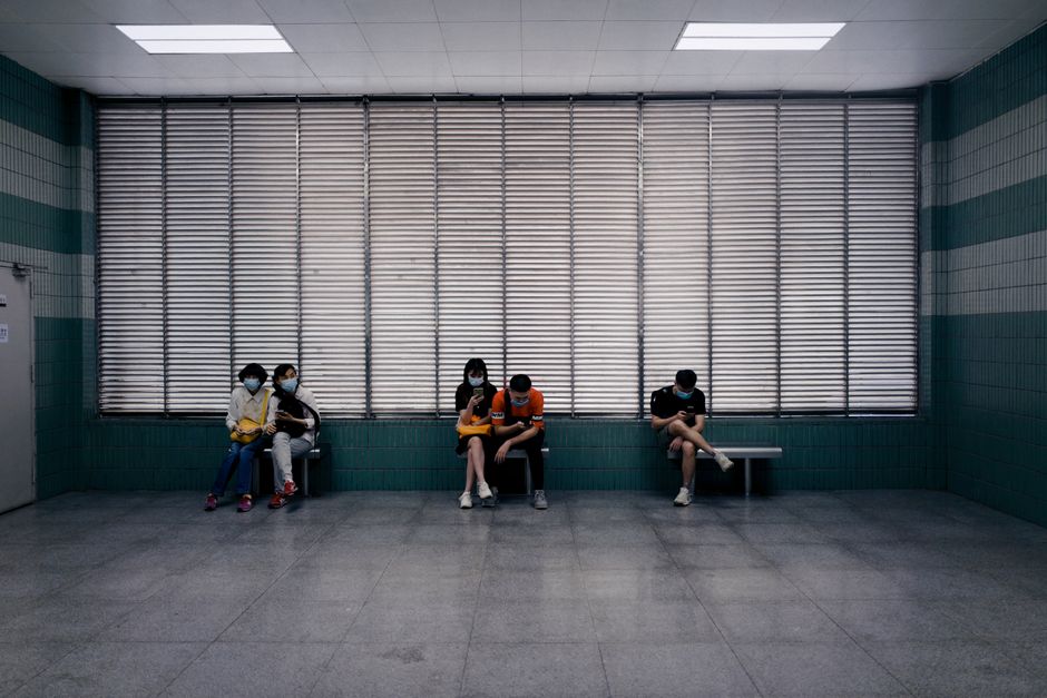 Full body of unrecognizable people in medical masks sitting on benches in spacious hall of clinic against windows