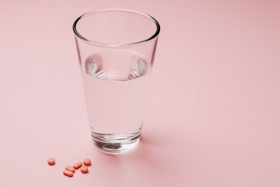 Glass of water with scattered pink pills on a pastel pink background, symbolizing medication or daily supplements.
