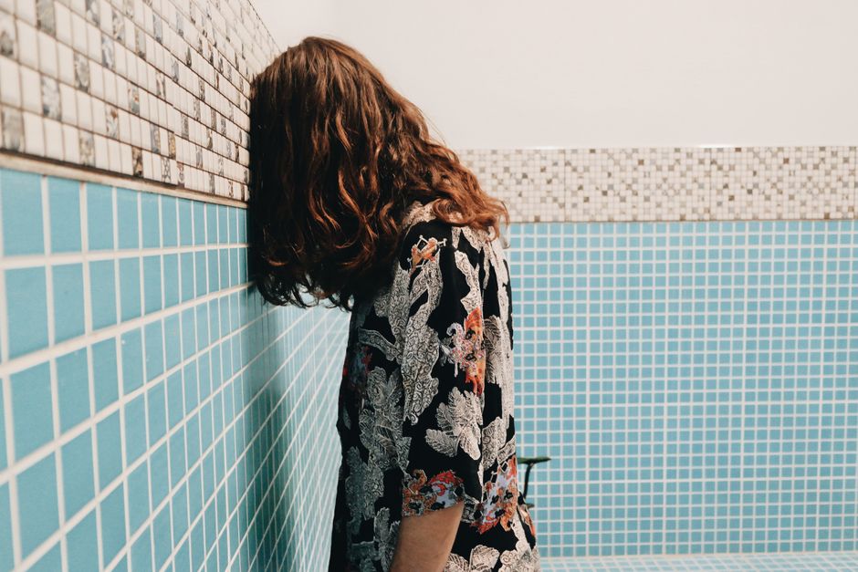 A person with long hair leans their head against a blue and mosaic tiled wall indoors.