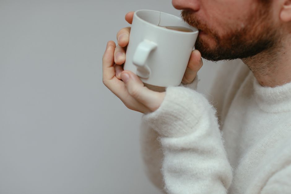 Close-up of a bearded man sipping a hot drink, wearing a cozy sweater indoors. Warm and inviting setting.