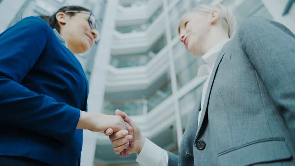 Two business professionals shaking hands in a modern office atrium, symbolizing collaboration.