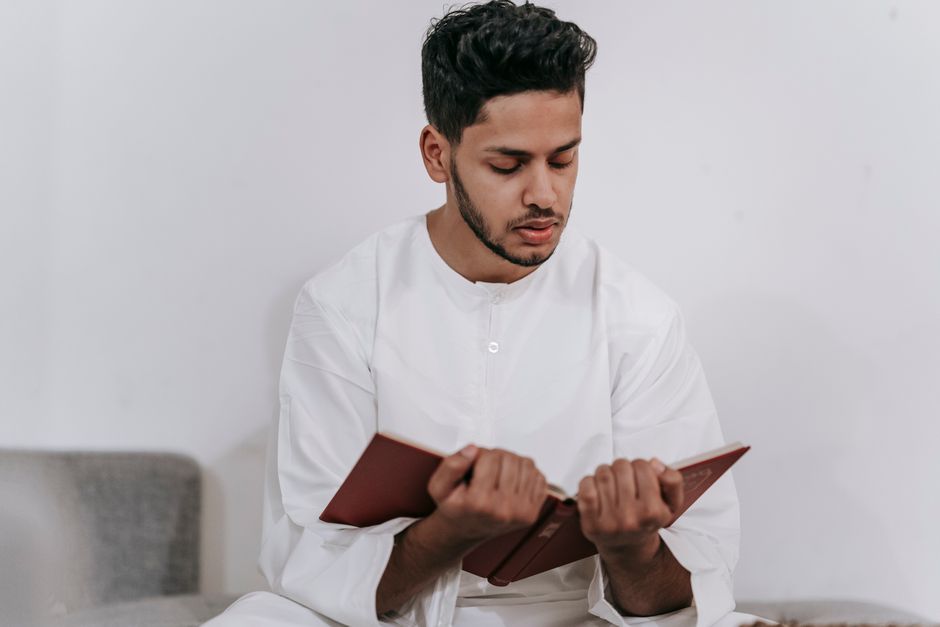 A young man in Islamic attire intimately reading the Quran indoors.