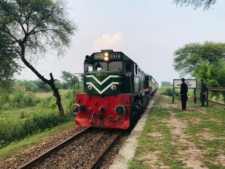 A locomotive passing through lush greenery with people waiting at a rural station.