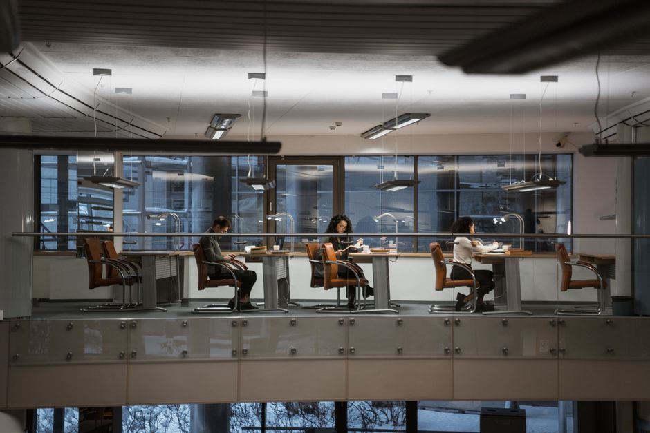 Adults studying in a modern library setting with large windows and ample lighting.