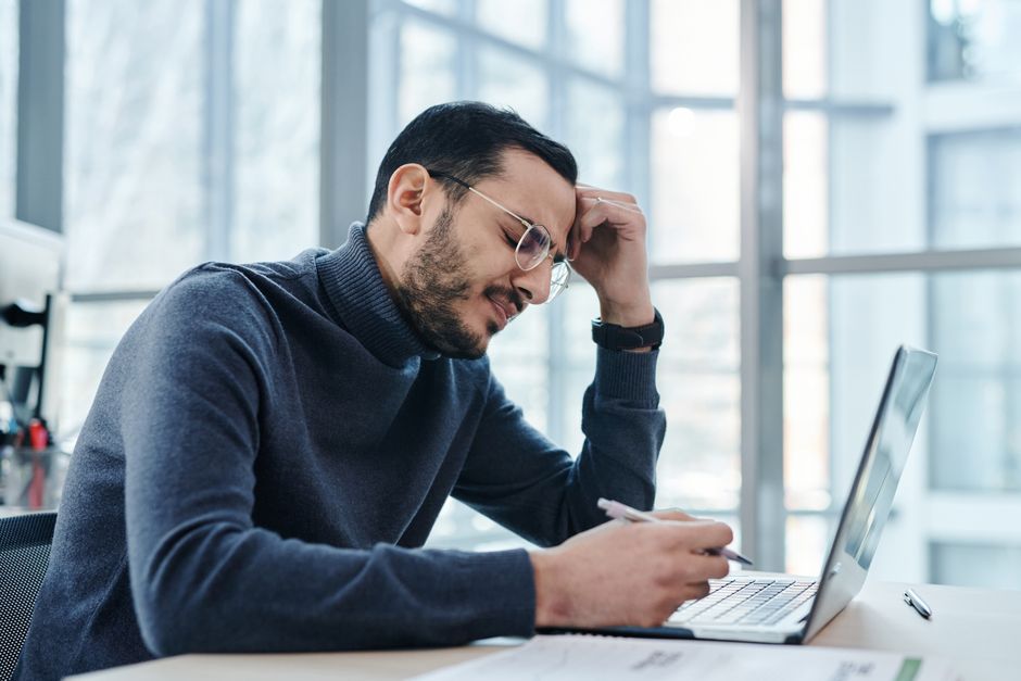 Man looking stressed at laptop in a modern office with large windows.