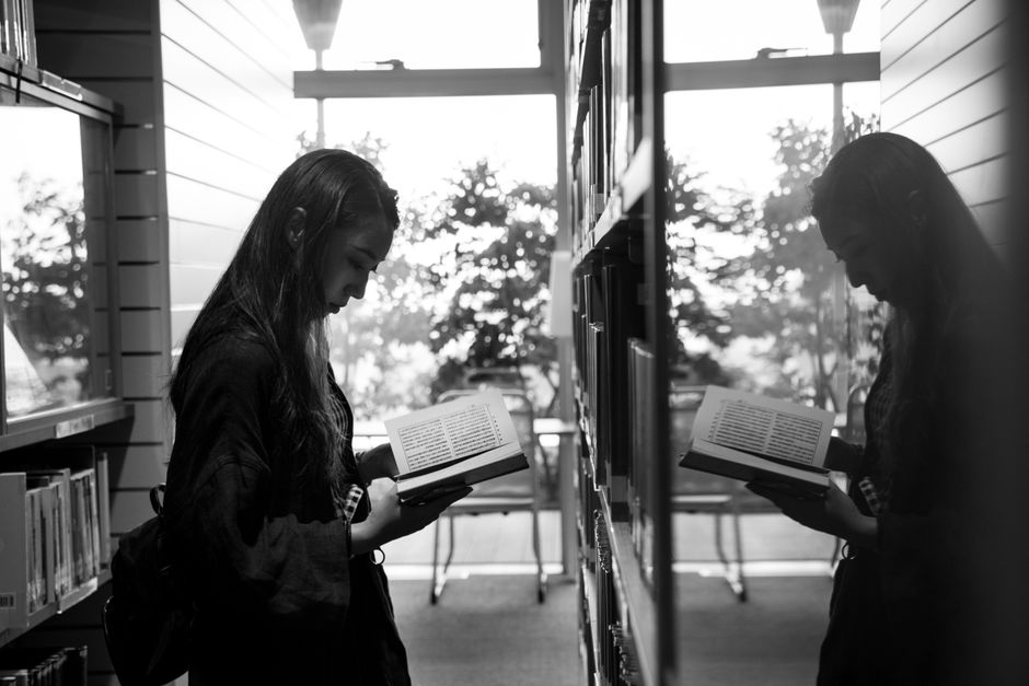 A woman engrossed in a book beside a reflective window in a library.