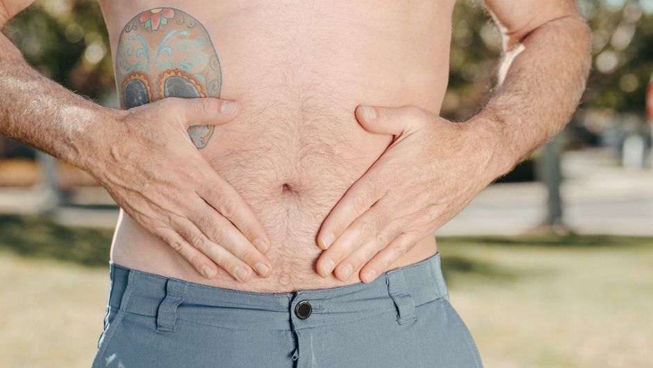 A man with a tattoo on his torso holds his stomach outdoors, suggesting discomfort.