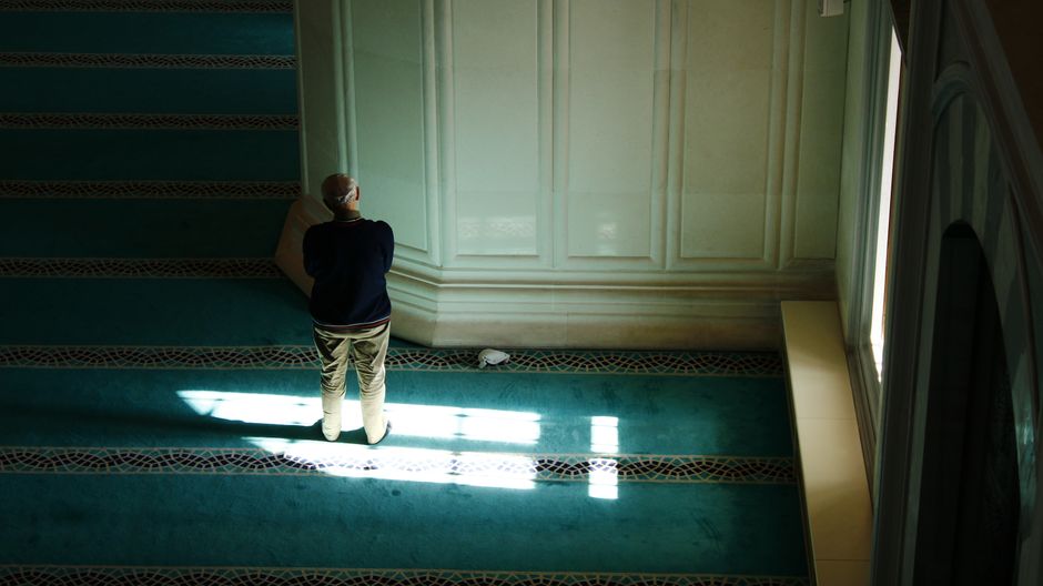 An adult stands in prayer in a sunlit mosque interior, creating a serene atmosphere.