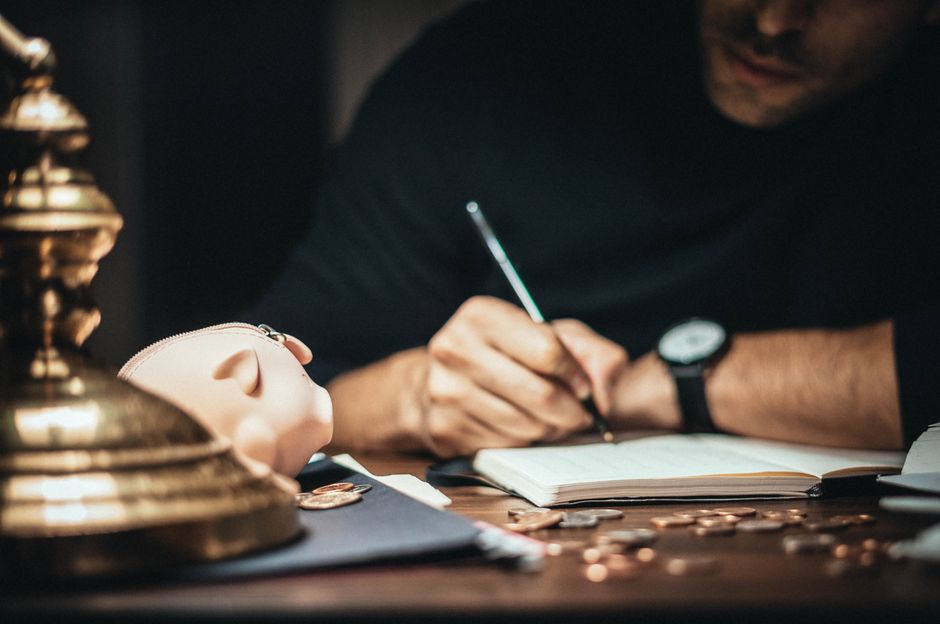 Crop man sitting at desk with vintage lamp and scattered coins and piggybank while writing in notepad