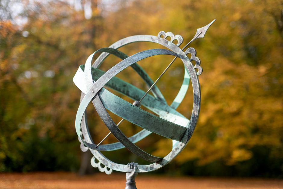An artistic sundial stands in focus with vibrant autumn leaves in the background, capturing a serene seasonal moment.
