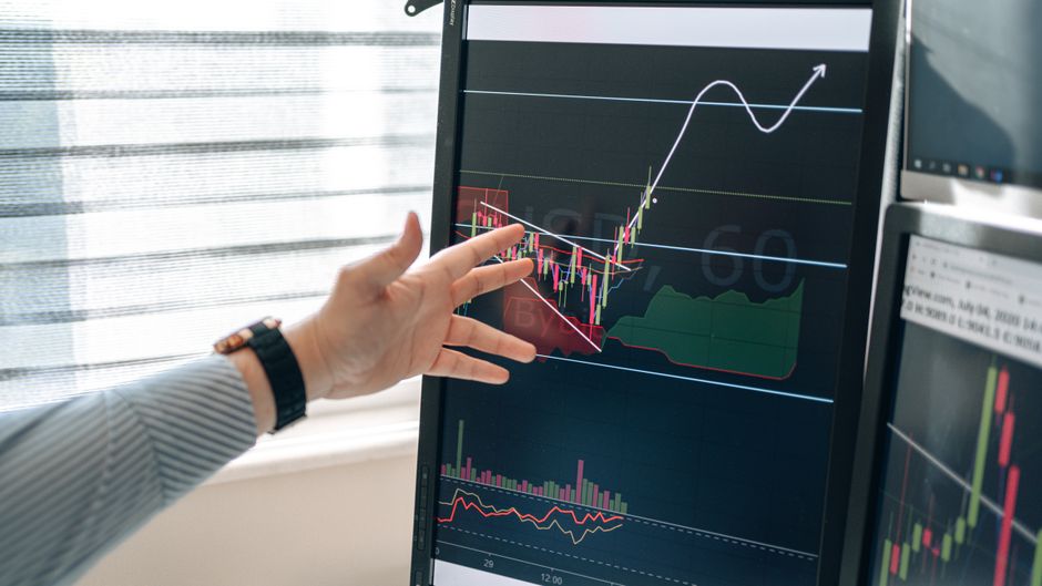 Close-up of a hand pointing at stock market graphs on a monitor in a workspace.