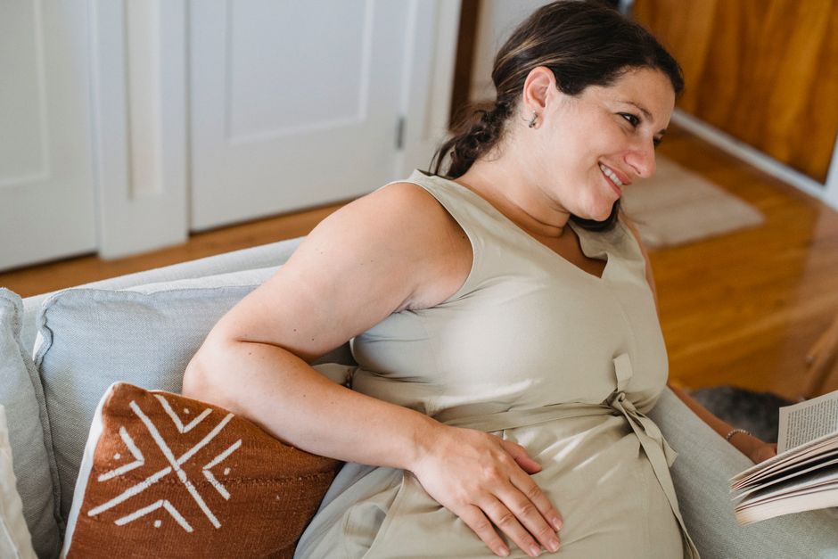 Young pregnant female in beige dress smiling and looking away while reading book on comfortable sofa