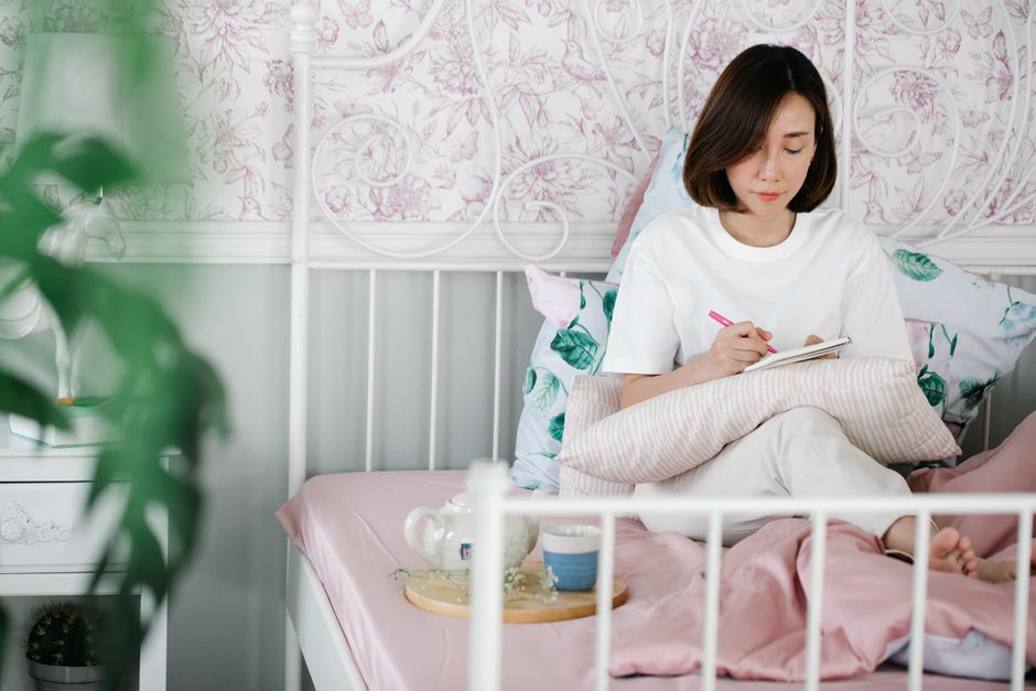 Mentel Health An Asian woman sitting on a bed, journaling in a serene and cozy bedroom environment.