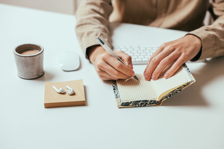 Person writing in a notebook on a desk with coffee and AirPods nearby.