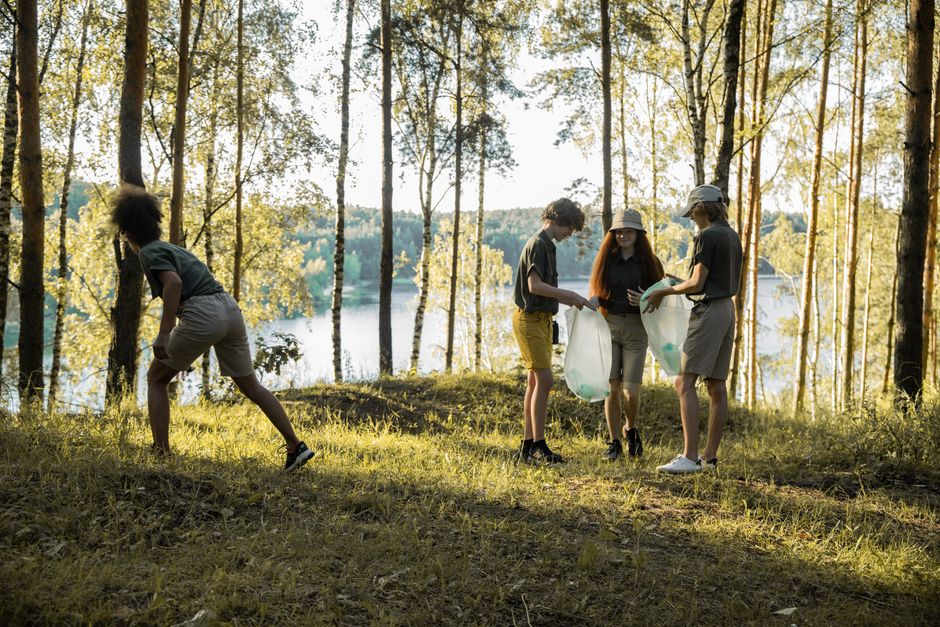 A group of teenagers clean up a forest by a lake, collecting trash in plastic bags.