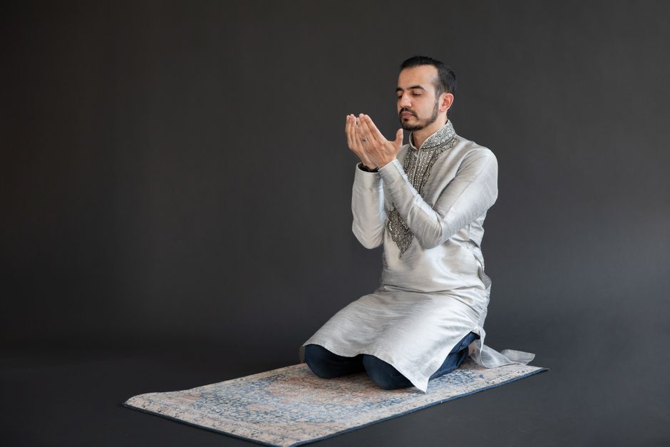 An adult man is kneeling and praying indoors on a prayer rug.