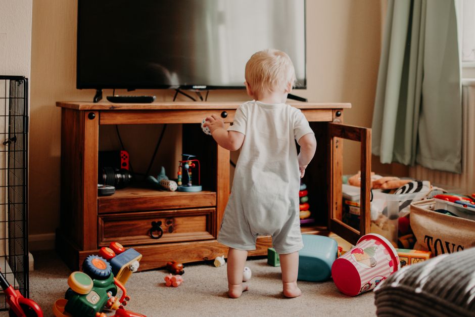 A toddler playing in a messy living room filled with colorful toys and a TV stand.