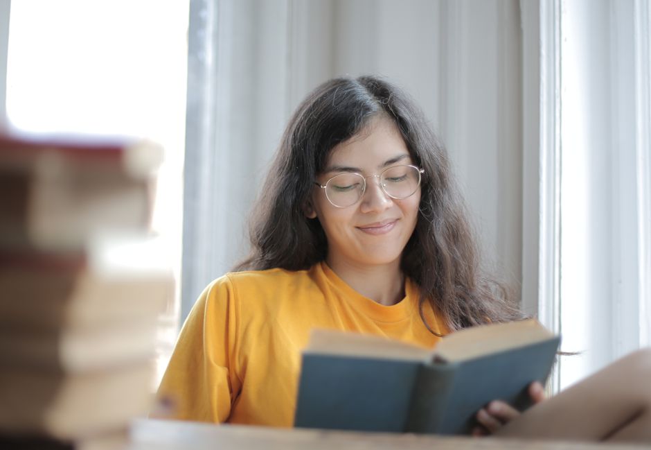 A smiling woman wearing a yellow shirt reads a book by a window indoors.