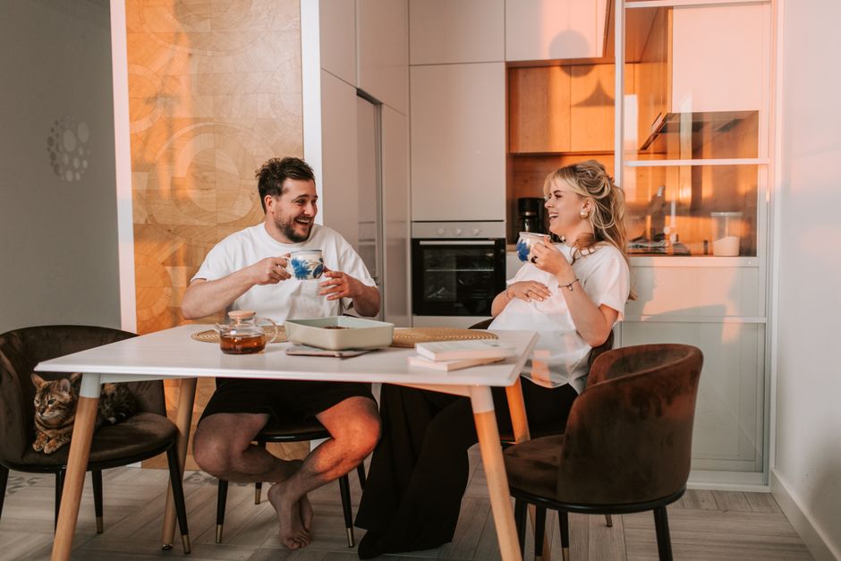 A happy pregnant couple sitting in a kitchen, enjoying tea and laughter together.