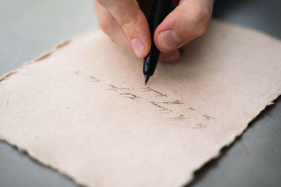 A close-up view of a person's hand writing with a black pen on textured paper.