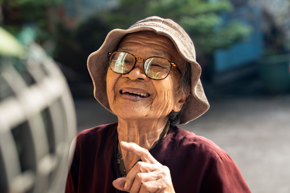 Elderly Vietnamese woman smiling joyfully outdoors in sunlight, wearing glasses and a sunhat.
