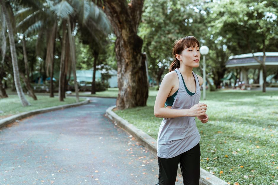 Thoughtful young Asian female athlete in sportswear running on asphalt road covered with faded leaves in city park in daytime and looking away