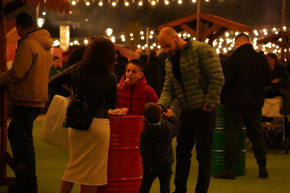 A family interacts at an evening street market with decorative lights and a bustling ambiance.