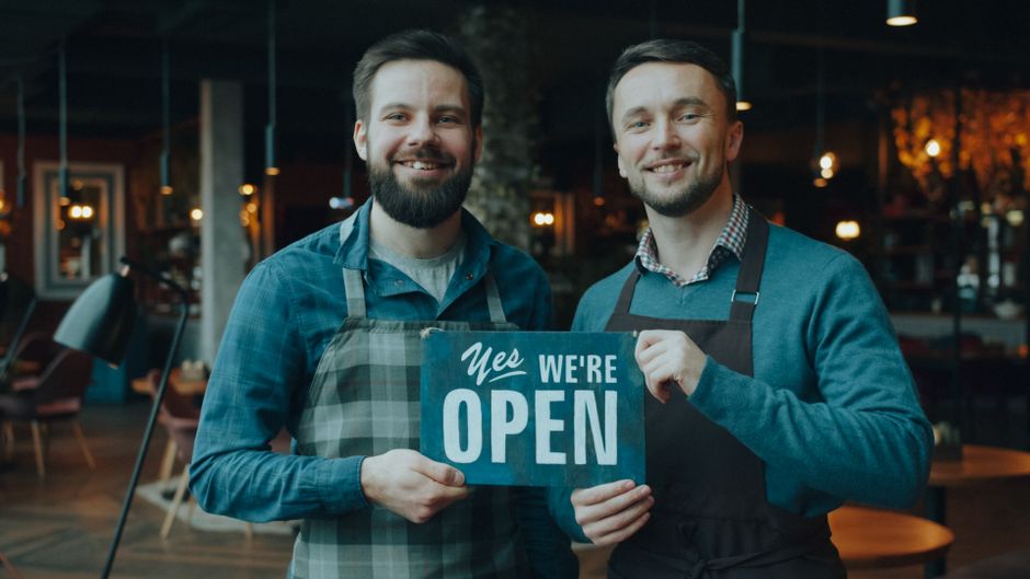Two cafe owners smiling and holding a 'Yes We're Open' sign inside their cozy cafe.