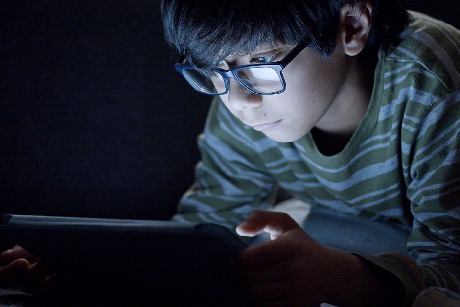 Young boy wearing glasses focused on tablet screen in dark room. Captures modern tech usage.